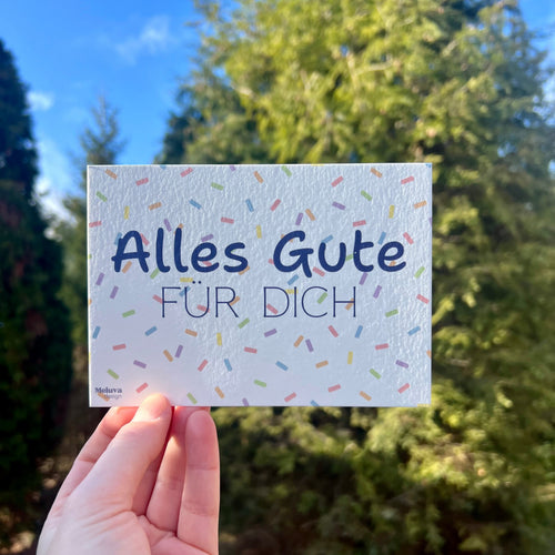 Eine Postkarte mit Konfetti-Hintergrund und dem Schriftzug Alles Gute fuer Dich" in blau vor einem grünen Strauch. 