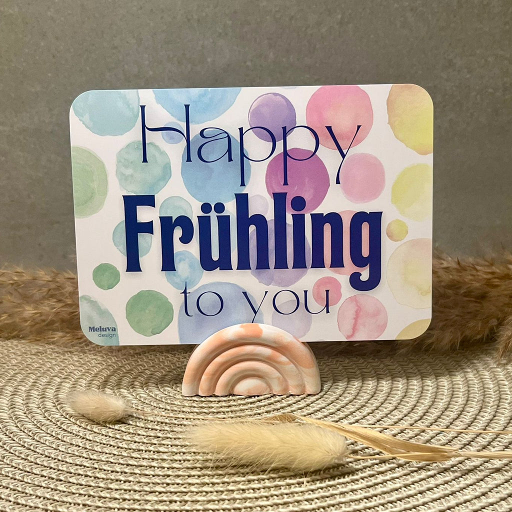 Eine Postkarte mit dem SchriftzugHappy Frühling to You in blau. Der Hintergrund besteht aus mehreren Kreisen in Bunten Farben (von links nach rechts ergibt sich ein Farbverlauf von grün über blau, lila, rot, orange zu gelb). Die Postkarte hat abgerundete Ecken. Die Postkarte befindet sich in einem orangenen Kartenhalter in Regenbogen-Form