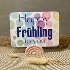 Eine Postkarte mit dem SchriftzugHappy Frühling to You in blau. Der Hintergrund besteht aus mehreren Kreisen in Bunten Farben (von links nach rechts ergibt sich ein Farbverlauf von grün über blau, lila, rot, orange zu gelb). Die Postkarte hat abgerundete Ecken. Die Postkarte befindet sich in einem orangenen Kartenhalter in Regenbogen-Form