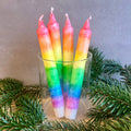 Ein Glas in dem vier Stabkerzen mit Regenbogen-Farbverlauf stehen. Um das Glas liegen Tannenzweige als Deko. 