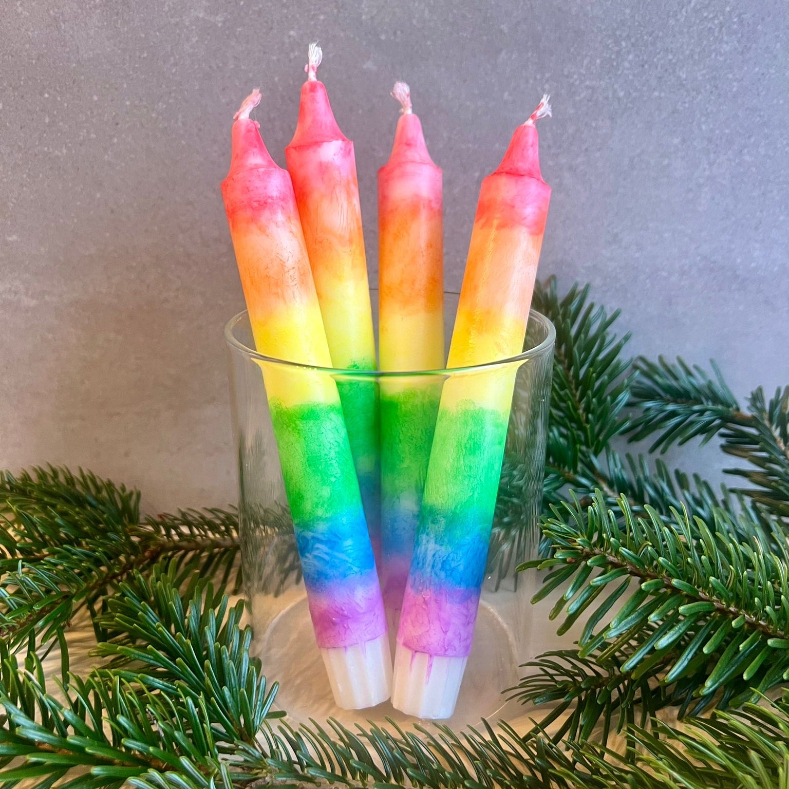 Ein Glas in dem vier Stabkerzen mit Regenbogen-Farbverlauf stehen. Um das Glas liegen Tannenzweige als Deko. 