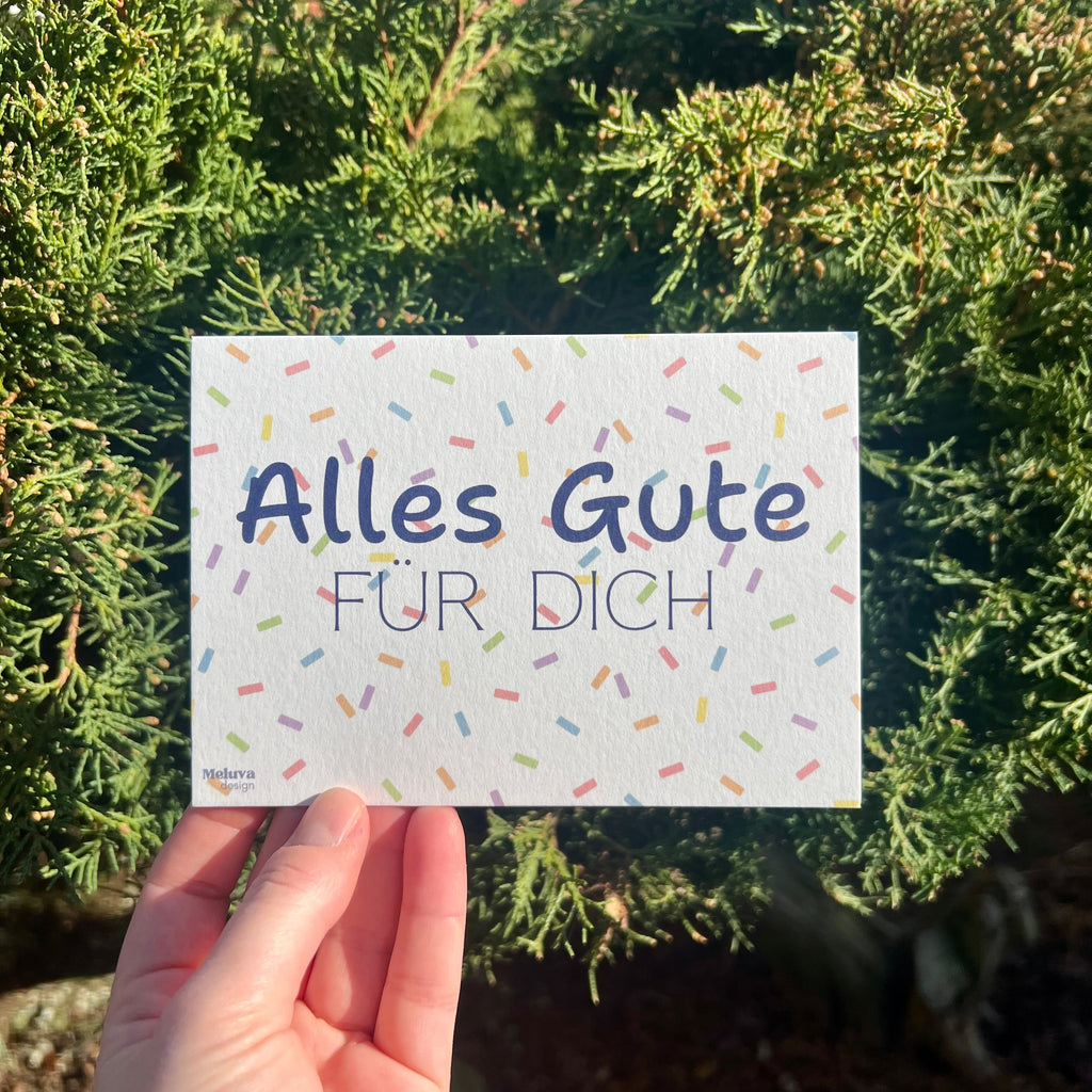 Eine Postkarte mit Konfetti-Hintergrund und dem Schriftzug Alles Gute fr Dich, die Postkarte wird vor einem grünen Strauch gehalten.