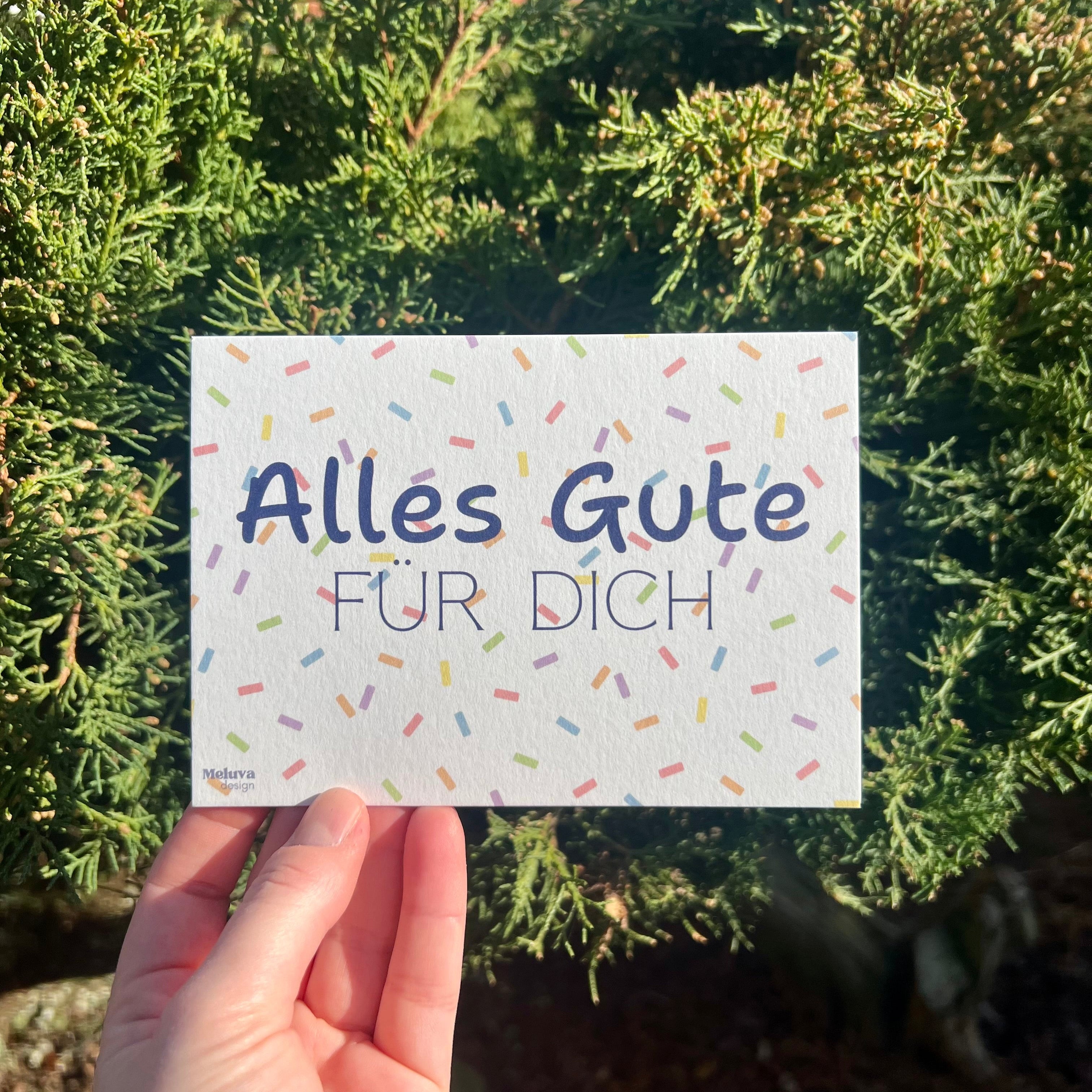 Eine Postkarte mit Konfetti-Hintergrund und dem Schriftzug Alles Gute fr Dich, die Postkarte wird vor einem grünen Strauch gehalten.