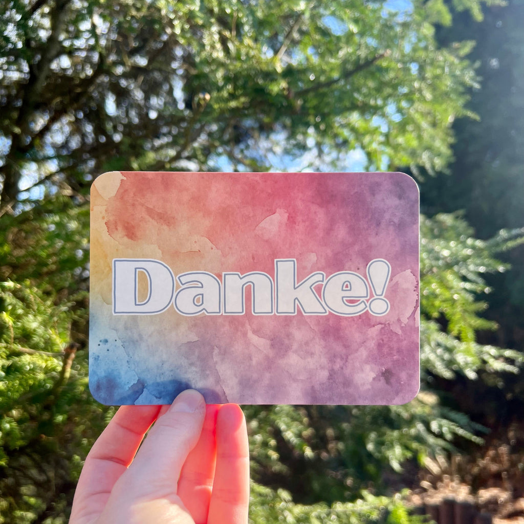 Eine Postkarte mit dem Schriftzug Danke mit abgerundeten Ecken, der Hintergrung ist ein bunter Verlauf von Aquarell-Farben, mit blau, gelb, orange und lila