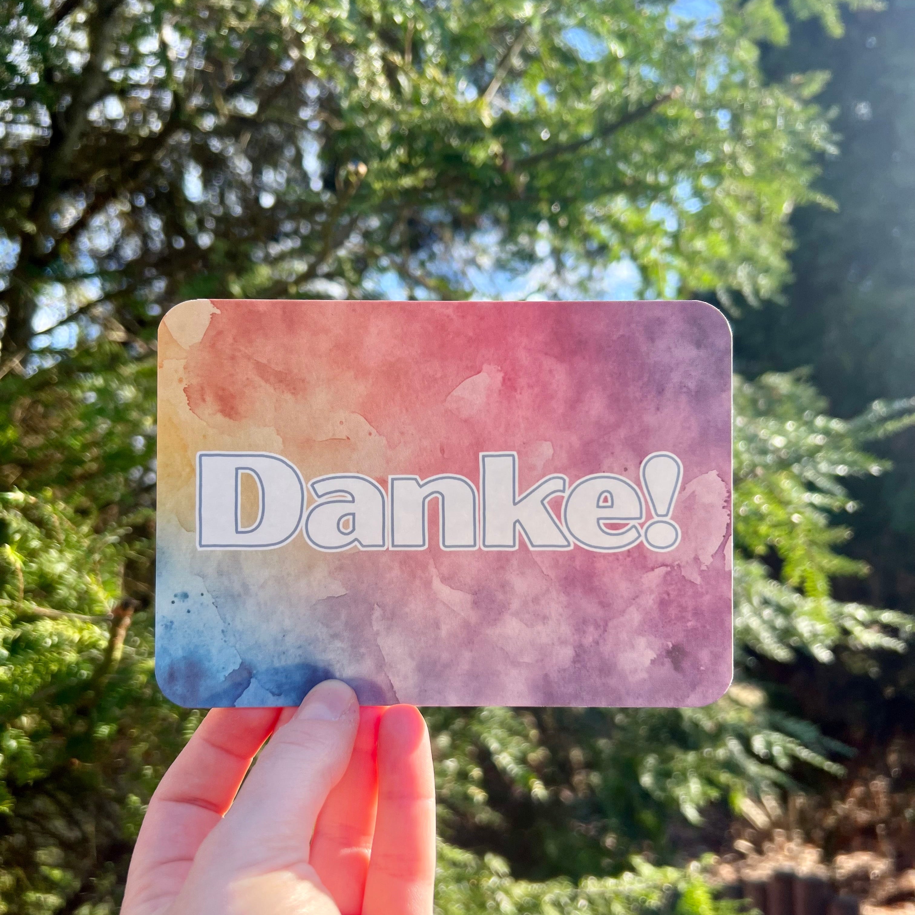 Eine Postkarte mit dem Schriftzug Danke mit abgerundeten Ecken, der Hintergrung ist ein bunter Verlauf von Aquarell-Farben, mit blau, gelb, orange und lila