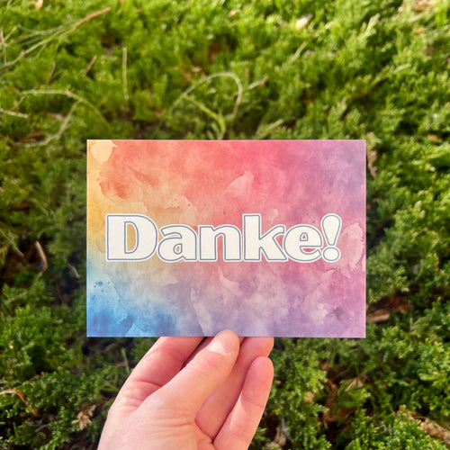 Eine Postkarte mit dem Schriftzug Danke mit abgerundeten Ecken, der Hintergrung ist ein bunter Verlauf von Aquarell-Farben, mit blau, gelb, orange und lila