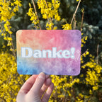 Eine Postkarte mit dem Schriftzug Danke mit abgerundeten Ecken, der Hintergrung ist ein bunter Verlauf von Aquarell-Farben, mit blau, gelb, orange und lila.