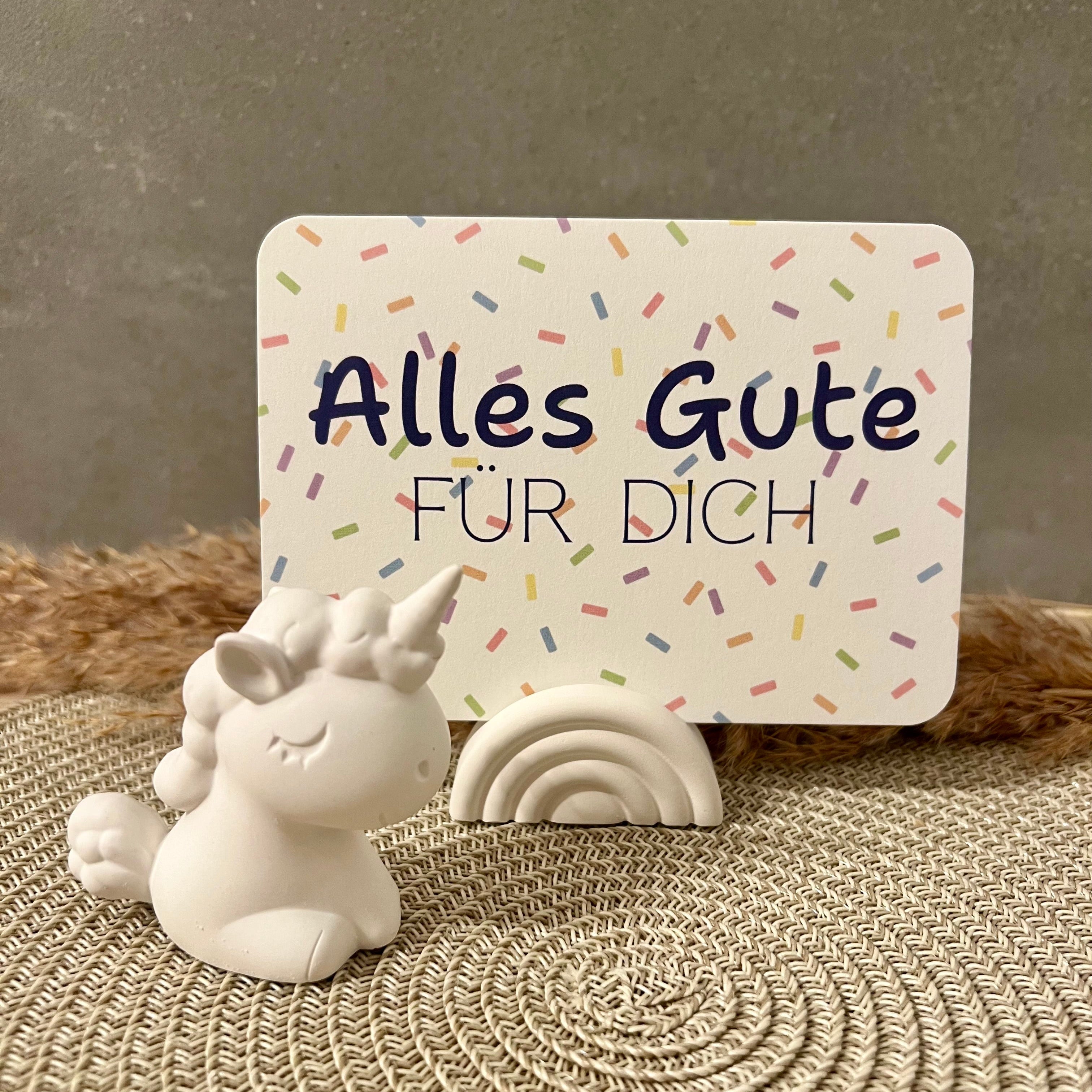 Eine Postkarte mit Konfetti-Hintergrund und dem Schriftzug "Alles Gute fr Dich" in blau steck in einem Regenbogen-förmigen Kartenhalter und daneben steht eine weiße Einhorn-Figur.