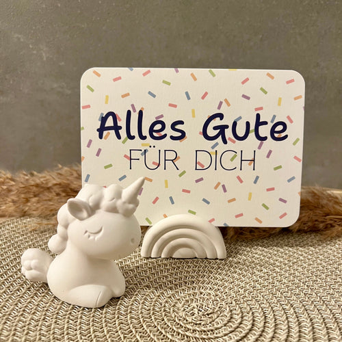 Eine Postkarte mit Konfetti-Hintergrund und dem Schriftzug "Alles Gute fr Dich" in blau steck in einem Regenbogen-förmigen Kartenhalter und daneben steht eine weiße Einhorn-Figur.