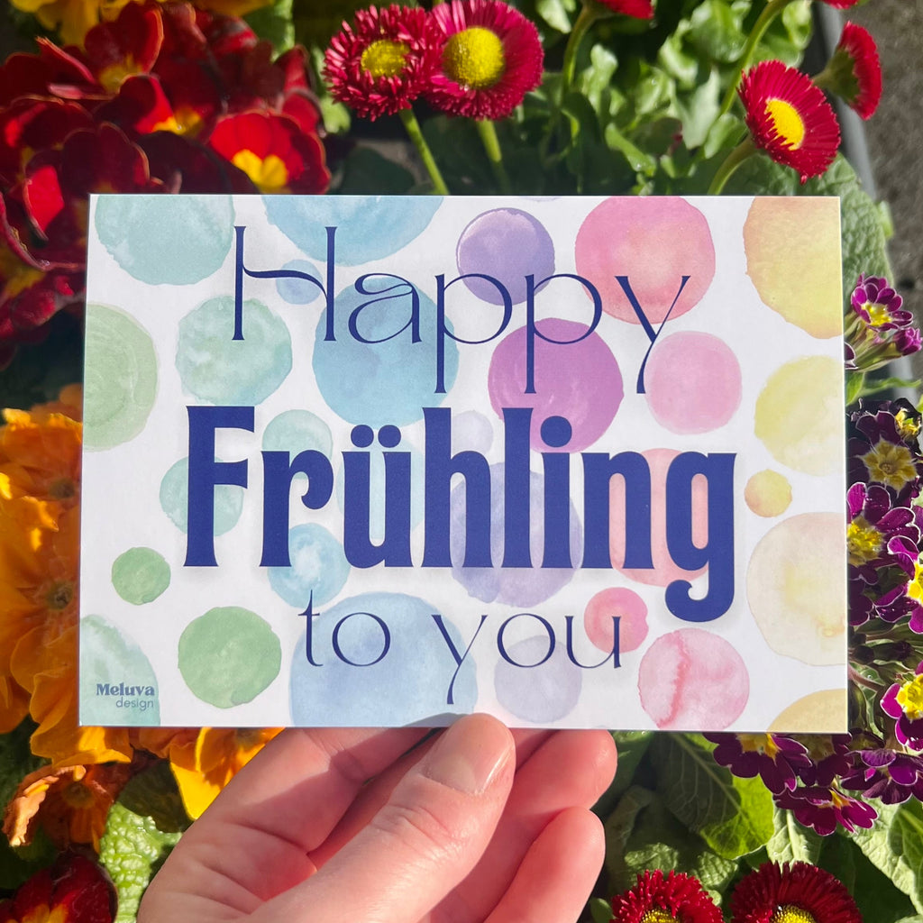 Eine Postkarte mit dem SchriftzugHappy Frühling to You in blau. Der Hintergrund besteht aus mehreren Kreisen in Bunten Farben (von links nach rechts ergibt sich ein Farbverlauf von grün über blau, lila, rot, orange zu gelb). Die Postkarte befindet sich vor mehreren bunten Blumen (rote und gelbe Primeln, rosa und weiße Gänseböümchen).