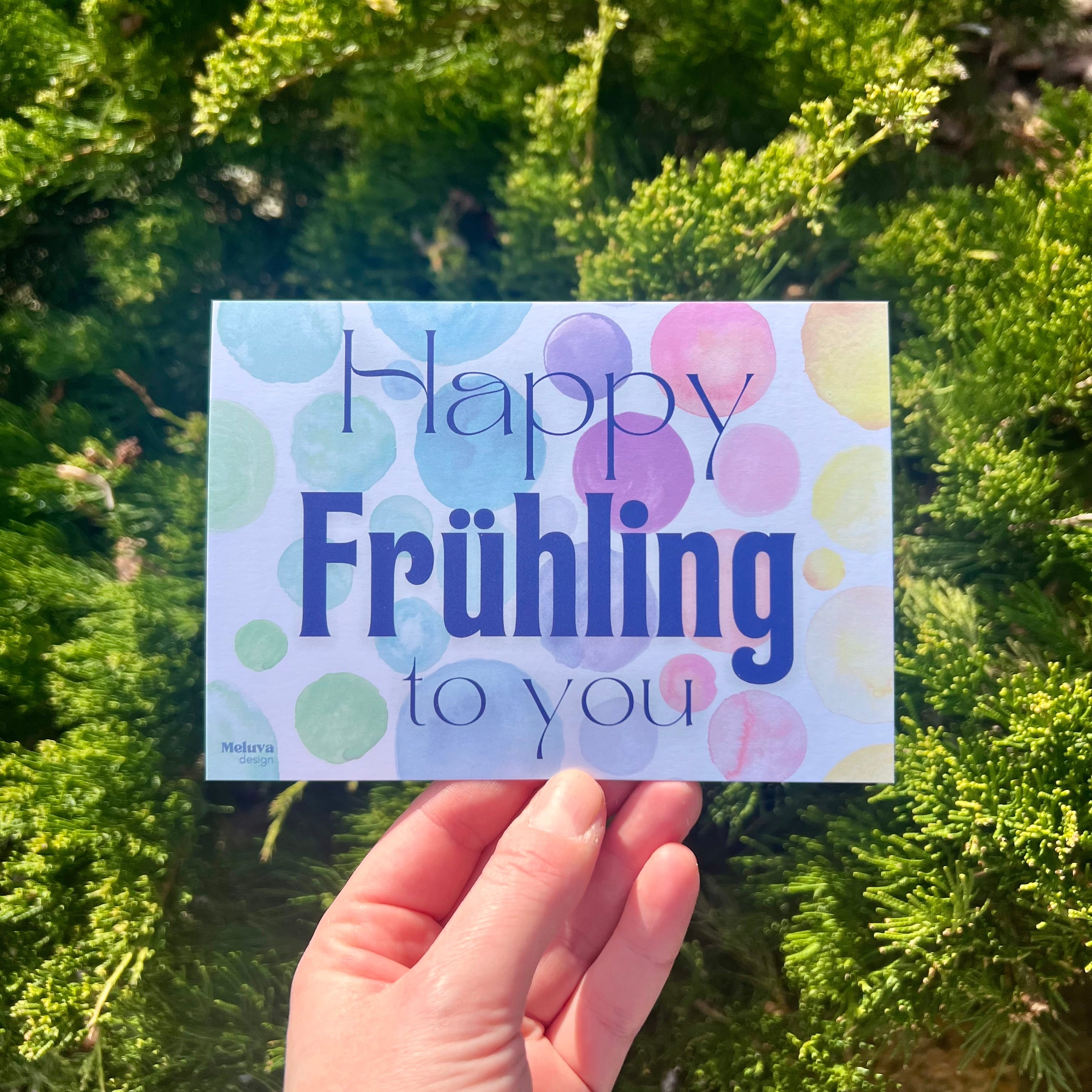Eine Postkarte mit dem SchriftzugHappy Frühling to You in blau. Der Hintergrund besteht aus mehreren Kreisen in Bunten Farben (von links nach rechts ergibt sich ein Farbverlauf von grün über blau, lila, rot, orange zu gelb). Die Postkarte hat eckige Ecken.  Die Postkarte wird vor einem Hintergrund aus einer grünen Pflanze mit Nadeln gehalten.