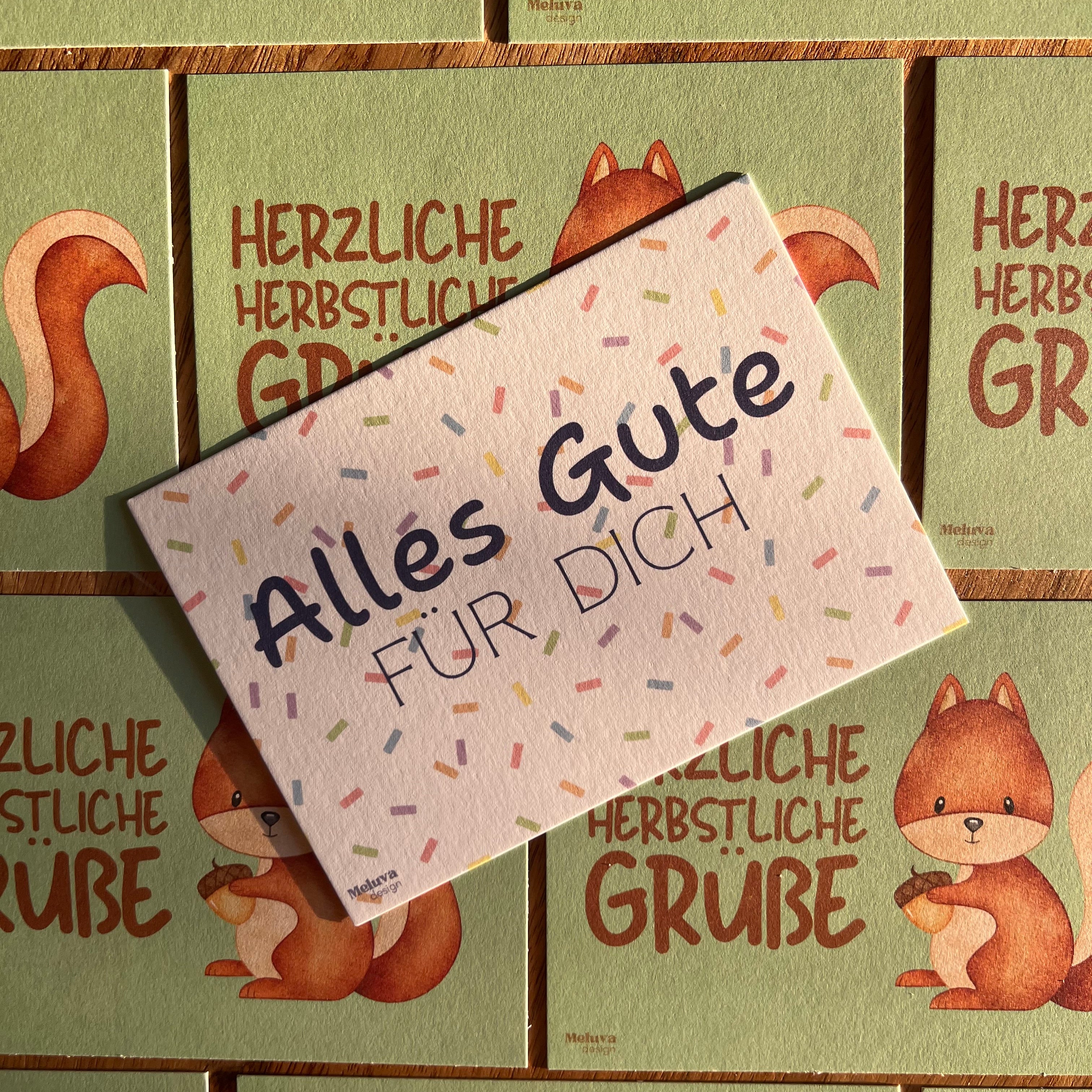 Eine Konfetti-Postkarte mit dem Schriftzug "Alles Gute für Dich" liegt auf einem Hintergrund von Postkarten mit einem Eichhörnchen und er Aufschrift "Herzliche Herbstliche Grüße"